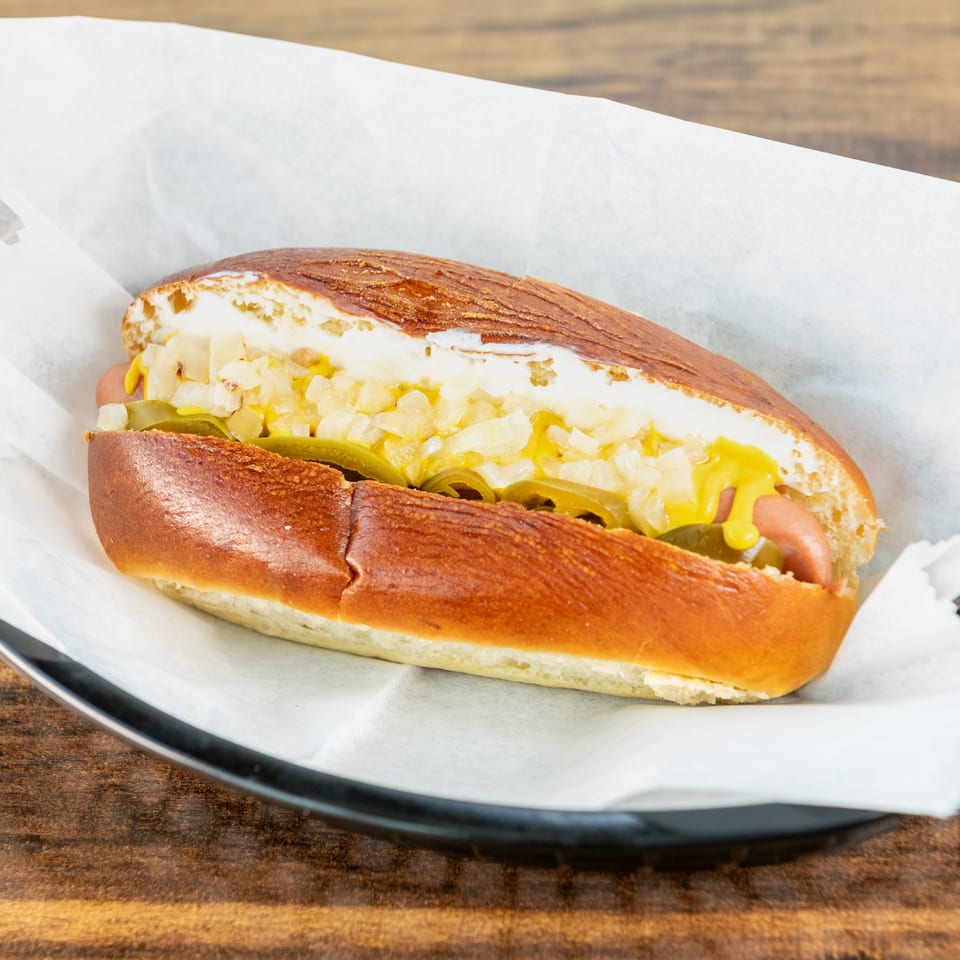 Hot dog in a toasted bun topped with mustard, diced onions, and sliced jalapeños on a paper-lined plate.