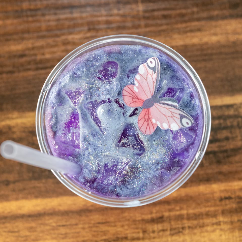 Purple glittery drink topped with a decorative pink butterfly in a clear glass on a wooden table.