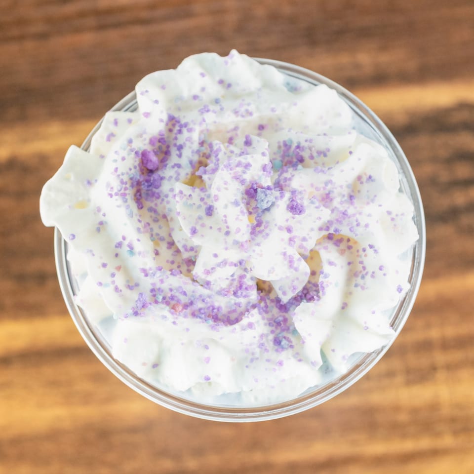 Overhead view of a glass dessert bowl filled with whipped cream, colorful sprinkles, and candy pieces on a wooden table.