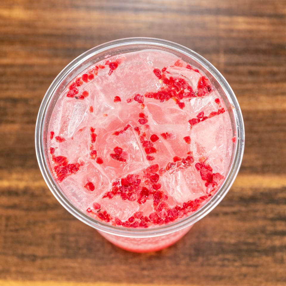 Pink iced beverage with crushed red berries in a clear glass on a wooden tabletop.