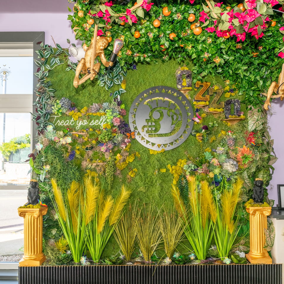 Lush restaurant greenery wall featuring succulents, flowers, golden monkeys, a circular logo, and the phrase “Treat yo self.”