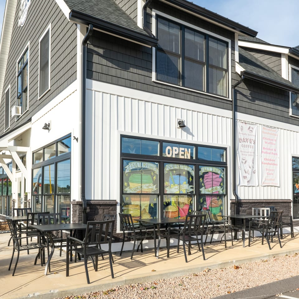 Dave’s Coffee shop exterior with outdoor seating, colorful illustrated windows, and banners advertising breakfast sandwiches, waffles, and barbecue.
